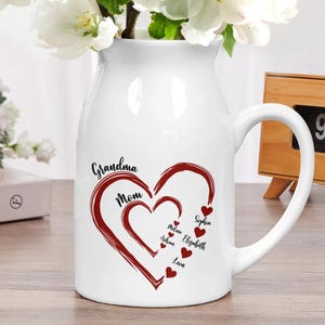 May include: White ceramic pitcher with a large handle, featuring a heart design in red with the words "Grandma" and "Mom". Smaller hearts with names are inside the larger heart. Flowers are in the pitcher.