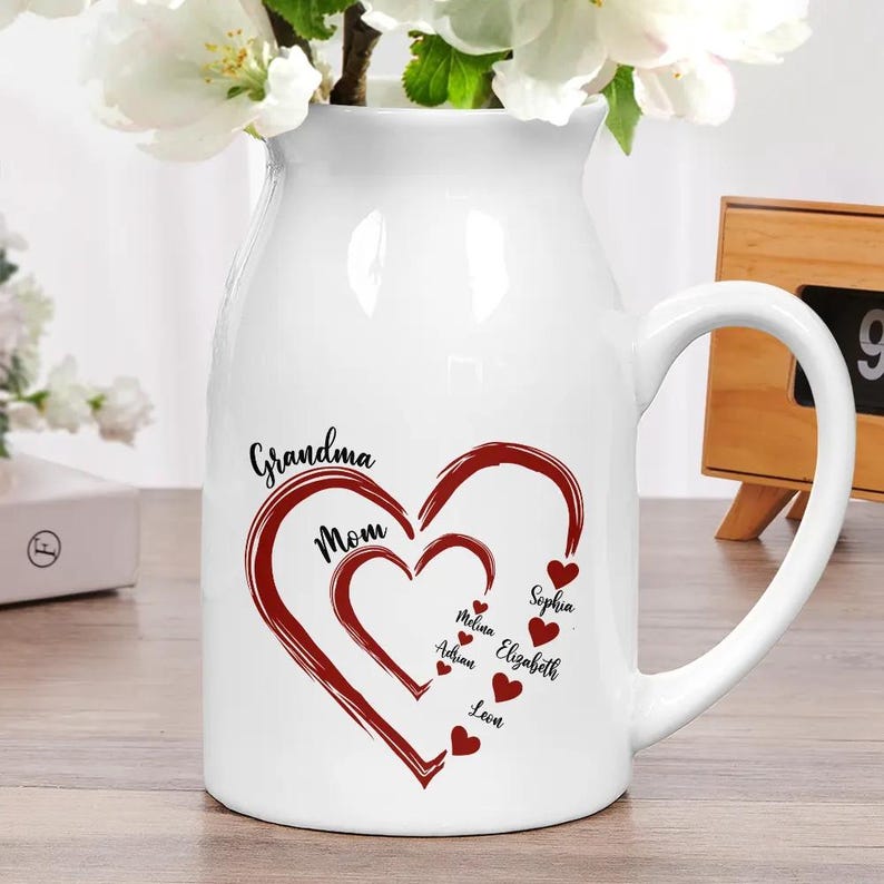 May include: White ceramic pitcher with a large handle, featuring a heart design in red with the words "Grandma" and "Mom". Smaller hearts with names are inside the larger heart. Flowers are in the pitcher.