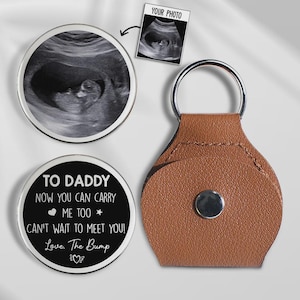 May include: A keychain featuring a brown leatherette holder and a silver ring. The keychain includes a round metal insert with a baby ultrasound image. Another insert has the text "TO DADDY NOW YOU CAN CARRY ME TOO! CAN'T WAIT TO MEET YOU! Love, The Bump."