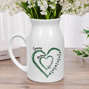 May include: White ceramic pitcher with a handle, filled with white flowers. The pitcher has a green heart design with the words "Gammy" and "Mommy" and names inside. A decorative vase for home decor.