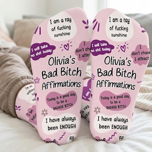 May include: A pair of light pink socks featuring purple and black text and graphics. The text reads: Olivia's Bad Bitch Affirmations, I am a ray of fucking sunshine, I will take no shit today, I don't chase I attract, Today is a good day to be a BADASS BITCH, and I have always been ENOUGH.