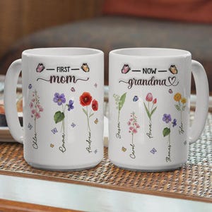 Op de afbeelding: Twee witte keramische mokken met bloemmotieven en de woorden "First Mom" en "Now Grandma". Elke mok heeft namen en kleurrijke bloemen zoals klaprozen, tulpen en viooltjes, met vlinderaccenten.