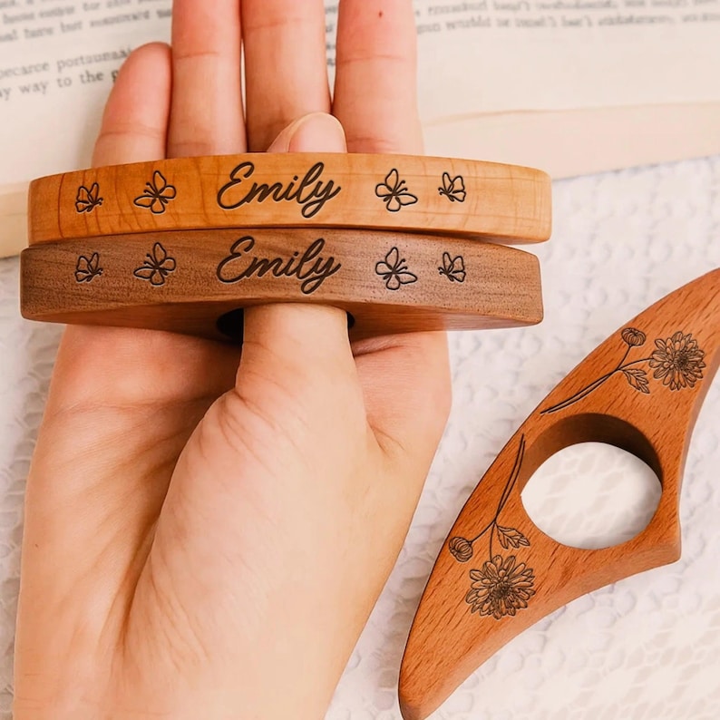 May include: Two wooden book page holders, one light and one dark, engraved with the name "Emily" and butterfly designs. A third wooden page holder with a floral design is also visible. These are perfect for book lovers.
