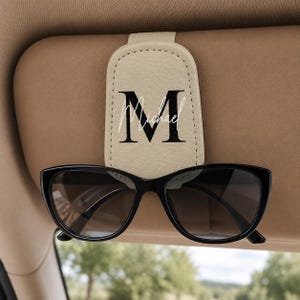 May include: A beige faux leather car visor sunglasses holder featuring a black capital letter M and the name Michael in white script text. The holder is attached to a tan car visor and holds a pair of black-framed sunglasses securely.