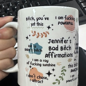 May include: White ceramic mug with black text and colorful graphics. The mug features phrases like "Bitch, you've got this" and "Jennifer's Bad Bitch Affirmations." Decorative elements include stars and floral designs. The mug has a white handle.