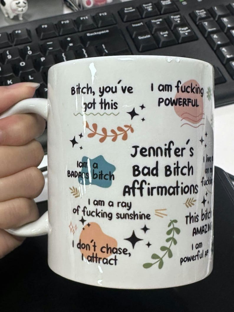 May include: White ceramic mug with black text and colorful graphics. The mug features phrases like "Bitch, you've got this" and "Jennifer's Bad Bitch Affirmations." Decorative elements include stars and floral designs. The mug has a white handle.