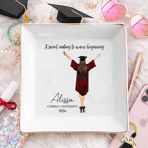 Könnte beinhalten: Eine weiße quadratische Keramik-Schmuckschale mit goldenem Rand. Die Schale zeigt eine Abschlussillustration einer Person in einem weinroten Kleid und Hut, mit dem Text "A sweet ending to a new beginning", dem Namen "Alissa", "Cornell University" und dem Jahr "2026".