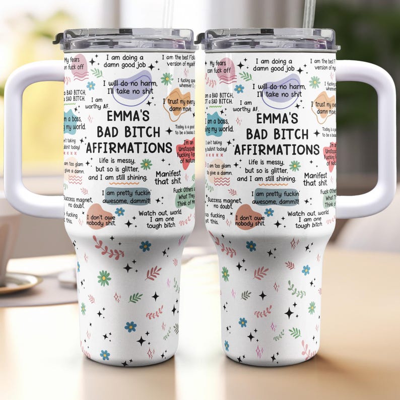 May include: Two white insulated tumblers with handles, featuring the text "EMMA'S BAD BITCH AFFIRMATIONS" and various affirmations. The tumblers have a white background with floral and star patterns, and black lids.