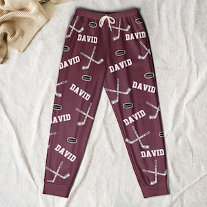 May include: Maroon lounge pants featuring a repeating pattern of white hockey sticks, black hockey pucks, and the name DAVID in white capital letters. The pants have an elastic waistband with a white drawstring and cuffed ankles.