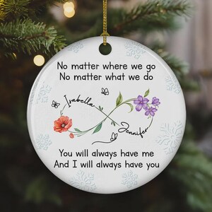 Op de afbeelding: Ronde witte keramische kerstversiering met een gouden ophangkoord. De versiering heeft de namen "Isabella" en "Jennifer" met bloemen- en vlinderaccenten. De tekst luidt "No matter where we go... You will always have me..."
