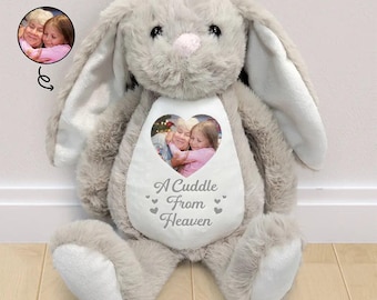 Op de afbeelding: Een grijze pluche konijnenknuffel met lange oren en een witte buikvlek. De buik is voorzien van een hartvormige foto van twee personen en de gedrukte tekst A Cuddle From Heaven, geaccentueerd met kleine grijze hartjes op de witte stof.