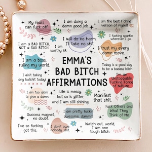 Può includere: Un vassoio in ceramica bianca con bordi dorati, con la scritta "EMMA'S BAD BITCH AFFIRMATIONS" in nero. Il vassoio è decorato con varie affermazioni in diversi caratteri e colori, tra cui frasi come "I am a boss" e "I'm an Unstoppable Fucking Force of Nature."