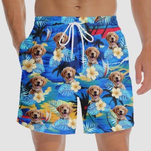 May include: Blue swim trunks with a tropical pattern featuring golden retriever puppies, flowers, palm trees, and sailboats. The trunks have a white drawstring and pockets.