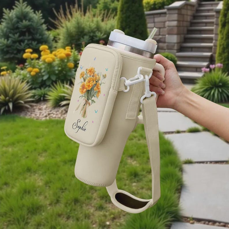 May include: A beige insulated water bottle with a matching carrying case and strap. The case features a floral bouquet design with the name "Sophie" printed below. The bottle has a silver lid and a clear straw.