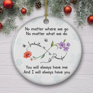 Op de afbeelding: Witte keramische kerstversiering met een gouden touwtje om op te hangen. De versiering heeft de tekst "No matter where we go, no matter what we do" en de namen "Isabella" en "Jennifer". Bloemen- en vlindermotieven zijn ook inbegrepen.