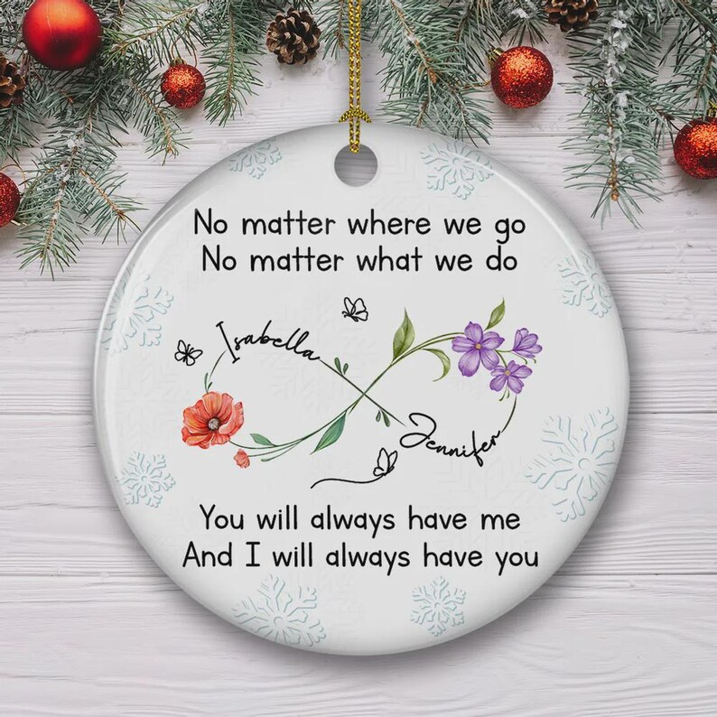 Op de afbeelding: Witte keramische kerstversiering met een gouden touwtje om op te hangen. De versiering heeft de tekst "No matter where we go, no matter what we do" en de namen "Isabella" en "Jennifer". Bloemen- en vlindermotieven zijn ook inbegrepen.