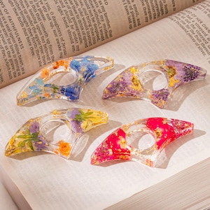 May include: Four clear resin thumb book page holders, each embedded with colorful dried flowers and gold flakes. The holders are in shades of blue, purple, yellow, and red, resting on an open book.