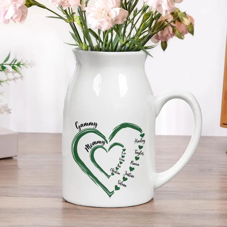 May include: White ceramic pitcher with a handle, featuring a green heart design with the words "Gammy" and "Mommy." Names like Hailey, Taylor, and others are listed within the heart. The pitcher holds pink flowers.