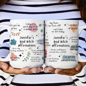 May include: Two white ceramic mugs with black text and colorful graphics. The mugs feature the words "Jennifer's Bad Bitch Affirmations" and various empowering phrases. The mugs are held by a person.