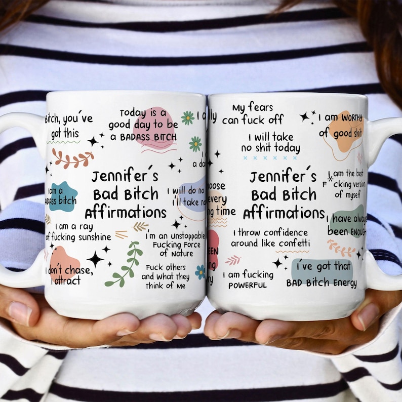 May include: Two white ceramic mugs with black text and colorful graphics. The mugs feature the words "Jennifer's Bad Bitch Affirmations" and various empowering phrases. The mugs are held by a person.