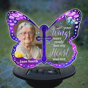 May include: A butterfly-shaped memorial garden stake with a photo of a woman, a cross, and the text "Your Wings were ready but my Heart was not." The stake has purple and blue accents and a solar-powered base.