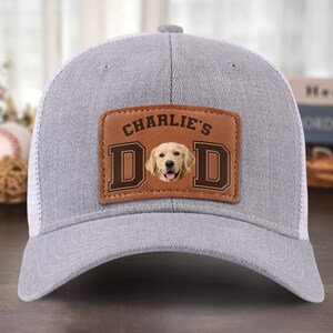 May include: Grey trucker cap with a white mesh back and a curved peak. The front has a brown leather patch with the text "CHARLIE'S DAD" and a photo of a golden retriever dog.