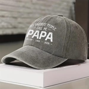 May include: A gray baseball cap with the text "MY FAVORITE PEOPLE CALL ME PAPA" in white. The names William, Linda, and Jason are printed below. The cap has a curved brim and a button on top.