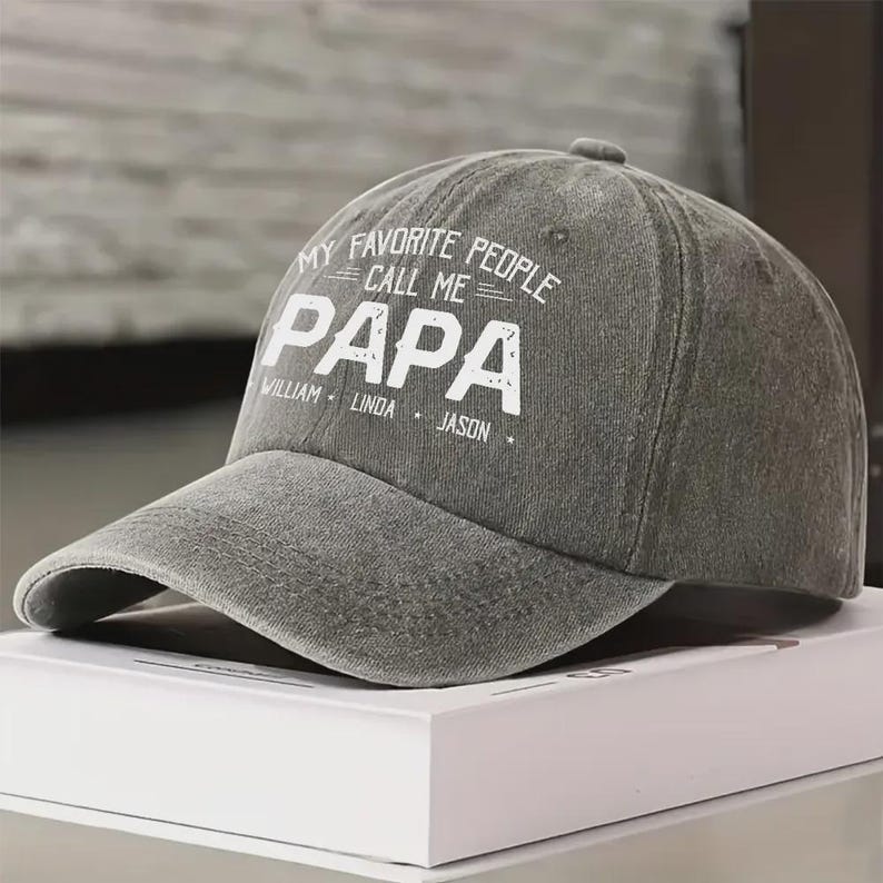 May include: A gray baseball cap with the text "MY FAVORITE PEOPLE CALL ME PAPA" in white. The names William, Linda, and Jason are printed below. The cap has a curved brim and a button on top.