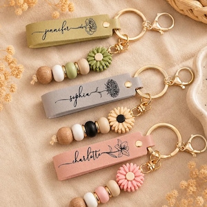 May include: Three personalized keychains with leather-like straps in olive green, gray, and dusty pink. Each strap features a name and a floral line drawing. Attached are gold-tone metal rings, lobster clasps, and strings of wooden and silicone beads.