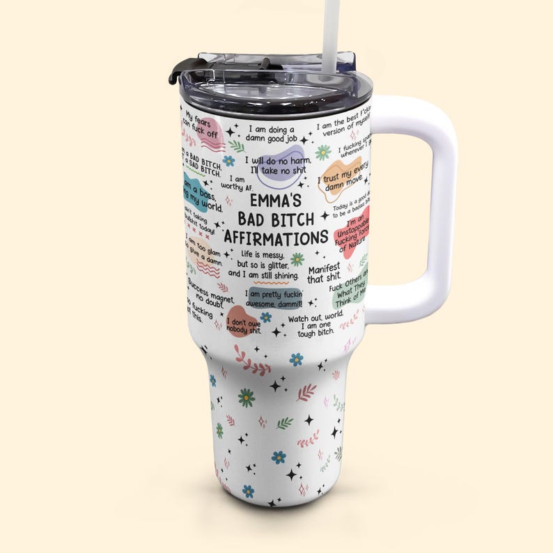 May include: A white tumbler with a handle and a straw, featuring the text "EMMA'S BAD BITCH AFFIRMATIONS" and various affirmations. The tumbler has a black lid and is decorated with floral and leaf patterns.