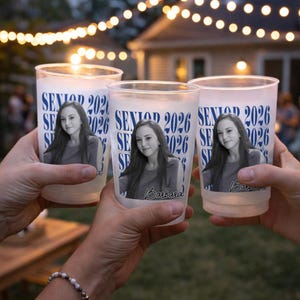 May include: Three frosted plastic cups with a photo of a young woman and the text "SENIOR 2026". The cups are held up in a toast, with string lights in the background. The name "Barbara" is printed on one of the cups.