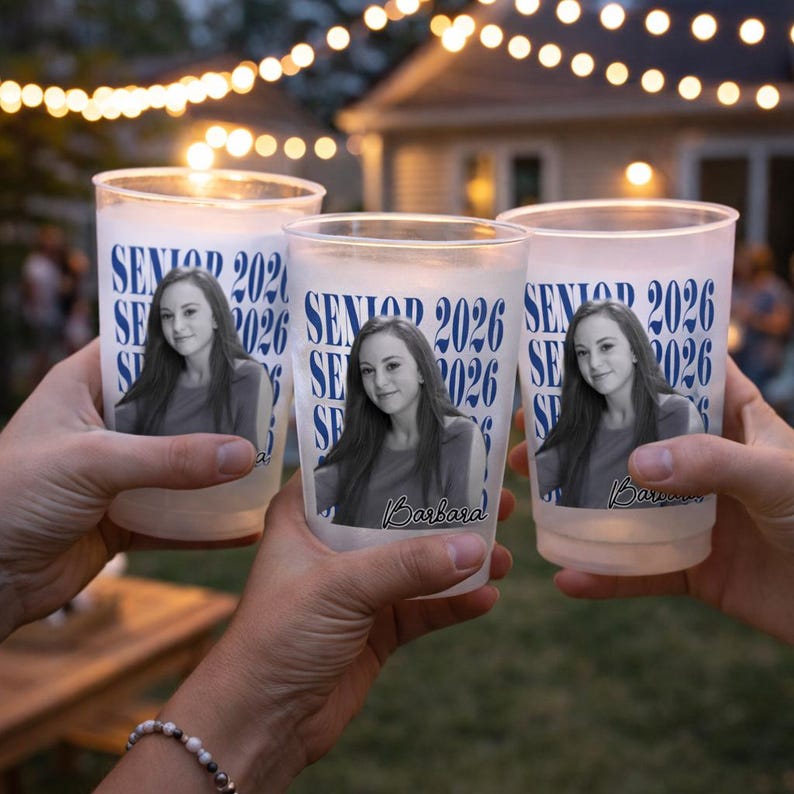 May include: Three frosted plastic cups with a photo of a young woman and the text "SENIOR 2026". The cups are held up in a toast, with string lights in the background. The name "Barbara" is printed on one of the cups.