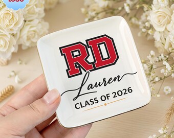 Op de afbeelding: Een wit keramisch sieradenschaaltje met gouden rand. Het schaaltje toont de letters "RD" in rood met een zwarte omlijning, de naam "Lauren" en "CLASS OF 2026" in zwart schrift. Het schaaltje is vierkant met afgeronde hoeken.
