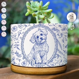 May include: A white ceramic cylindrical planter featuring a blue line-art illustration of a small dog inside an oval frame, with the name Charlie written below. The pot is decorated with a blue floral pattern and sits on a round wooden base.