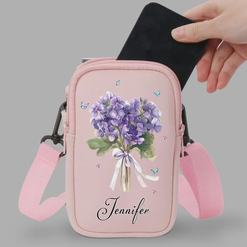 May include: A pink crossbody bag with a floral design and the name "Jennifer" printed on the front. The bag features a bouquet of purple flowers, butterflies, and a pink strap. A black smartphone is being placed inside the bag.