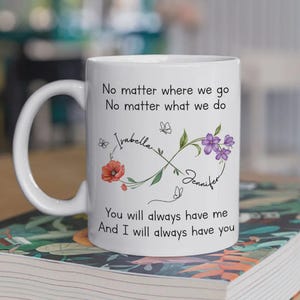 Op de afbeelding: Witte keramische mok met een gebogen handvat. De mok heeft de tekst "No matter where we go, no matter what we do, you will always have me, and I will always have you." Bloemen- en vlinderontwerpen met de namen "Isabella" en "Jennifer" zijn ook aanwezig.