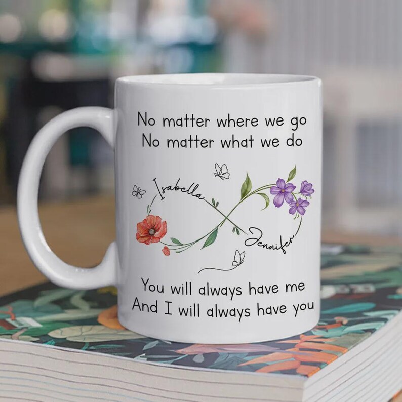 Op de afbeelding: Witte keramische mok met een gebogen handvat. De mok heeft de tekst "No matter where we go, no matter what we do, you will always have me, and I will always have you." Bloemen- en vlinderontwerpen met de namen "Isabella" en "Jennifer" zijn ook aanwezig.