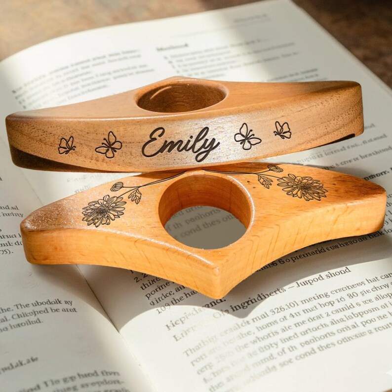 May include: Two wooden thumb page holders, one with the name "Emily" and butterfly designs, the other with a floral pattern. The thumb rests are on an open book, showcasing their use for reading.