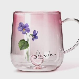 May include: A clear glass mug with a pink ombre effect. The mug features a watercolor design of purple violets and the name "Linda" in cursive. A heart-shaped base adds a decorative touch to this personalized drinkware.