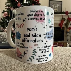 May include: White ceramic mug with the text "Pam's Bad Bitch Affirmations" and various affirmations in black, pink, and blue. The mug features floral and star designs, with a white handle.
