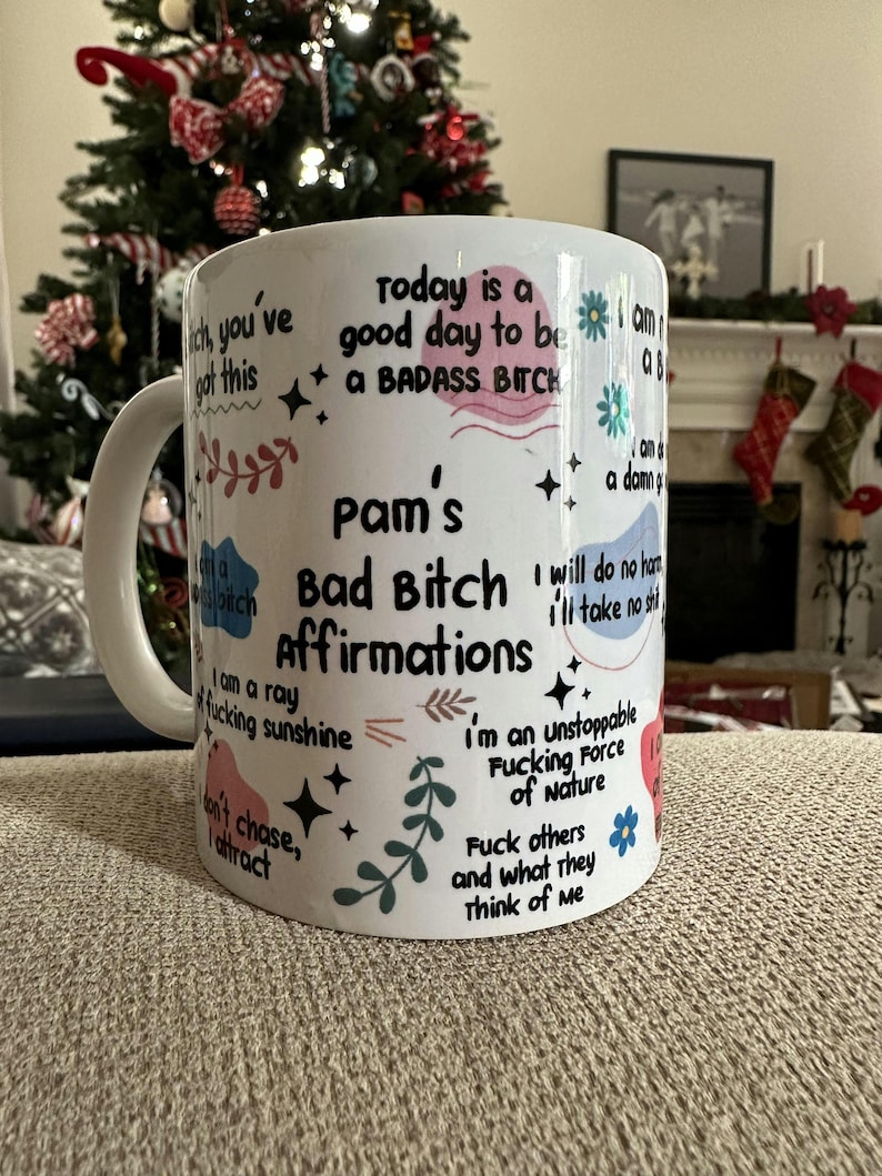 May include: White ceramic mug with the text "Pam's Bad Bitch Affirmations" and various affirmations in black, pink, and blue. The mug features floral and star designs, with a white handle.