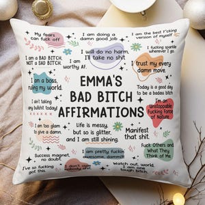 May include: A white square cushion printed with the words "EMMA'S BAD BITCH AFFIRMATIONS" in black. The design includes various positive affirmations in different fonts and colours, such as "I am worthy AF."