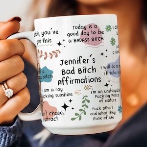 May include: White ceramic mug with the text "Jennifer's Bad Bitch Affirmations" and various affirmations in black, pink, and green. The mug features floral and star designs. The mug is held by a person.