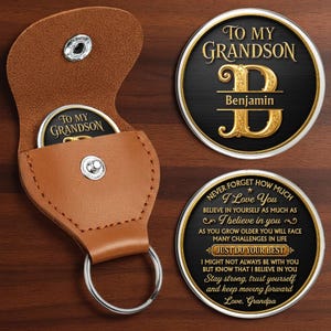 Peut inclure: Une pochette en cuir beige avec un bouton-pression argenté et un porte-clés, contenant une pièce ronde avec le texte TO MY GRANDSON. Deux pièces rondes supplémentaires sont visibles, l'une avec une lettre B dorée et Benjamin, l'autre avec du texte.