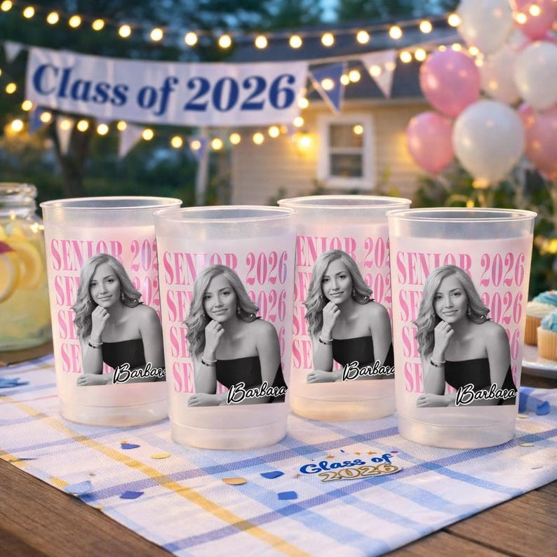 May include: Four frosted plastic cups with a pink "Senior 2026" design and a photo of a woman in a black dress. The cups are on a checkered tablecloth with a "Class of 2026" banner in the background.