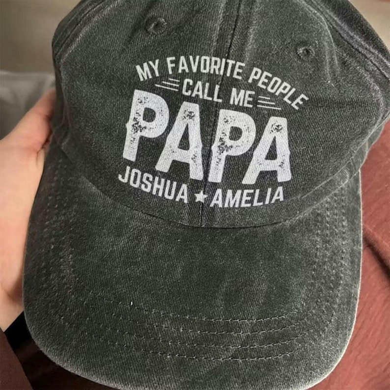 May include: A dark gray baseball cap with the text "MY FAVORITE PEOPLE CALL ME PAPA" in white. Below the word "PAPA" is the text "JOSHUA * AMELIA". The cap has a curved brim and a fabric strap.