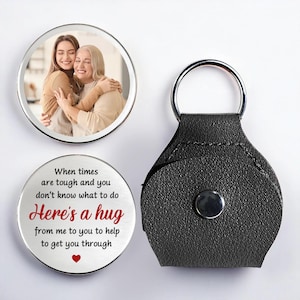 May include: A silver keyring with a black leather holder and two round metal charms. One charm displays a photo of two people embracing, the other has the text "Here's a hug" in red and black.