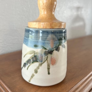 Puede incluir: Un tarro de cerámica con tapa de madera. El tarro es blanco con diseños florales azules y verdes. La tapa es de color marrón claro. El tarro está sobre una superficie de madera.