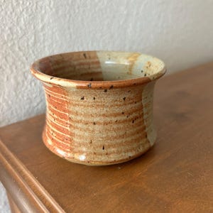 May include: A handmade ceramic bowl with a wide, slightly flared rim. The bowl has a speckled, earthy colour palette with bands of tan, orange, and brown. The interior is a lighter tan colour. The bowl sits on a wooden surface.