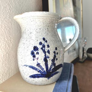 May include: A white ceramic pitcher with a blue floral design. The pitcher has a speckled glaze and a rounded body with a handle.